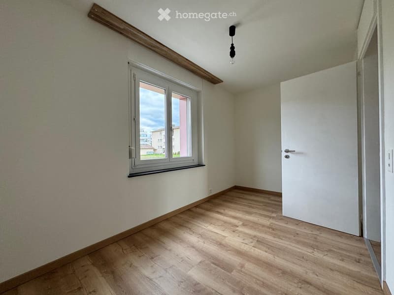 Empty room with wooden flooring, white walls, a window with a view, and a closed white door.