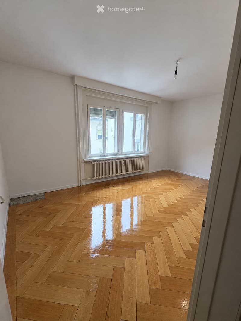 Empty room with polished wooden herringbone flooring, white walls, large window with blinds, radiator, and a single light fixture on the ceiling.