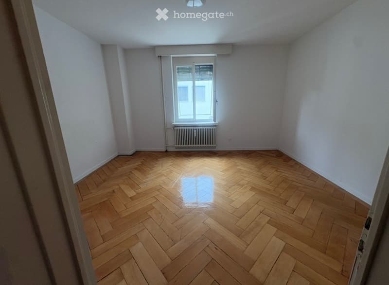 Empty room with wooden herringbone flooring, white walls, and a window with blinds. No furniture or equipment present.