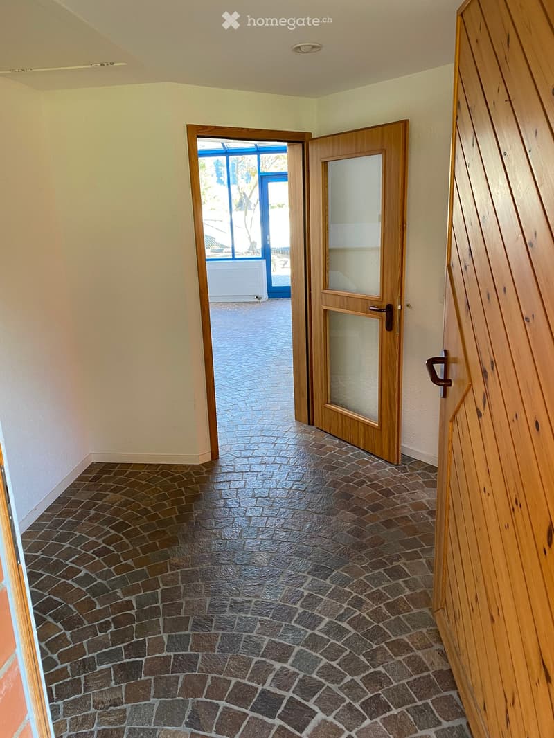 Empty hallway with tiled floor, wooden doors, and a view to an outdoor area through an open doorway.