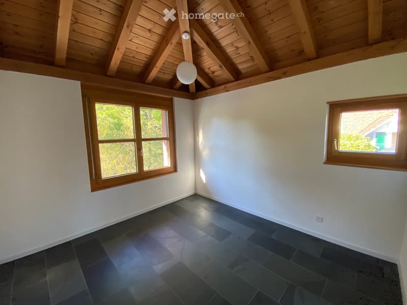 Empty room with wooden slanted ceiling, dark tiled floor, and large windows with wooden frames, allowing natural light to enter.