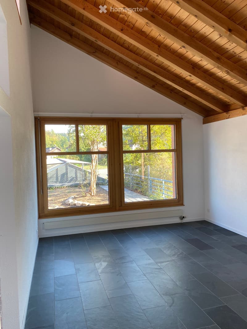 Empty room with wooden slanted ceiling, large window with wooden frame, grey tiled floor, white walls, radiator under window, and visible outdoor area with trees and fence.