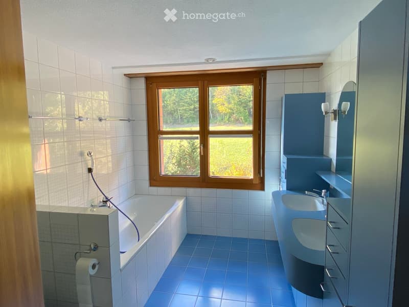 Modern bathroom with a bathtub, double sink, mirror, and blue cabinetry. The room features white tiled walls, blue tiled flooring, and a large window with wooden frames offering a view of greenery.
