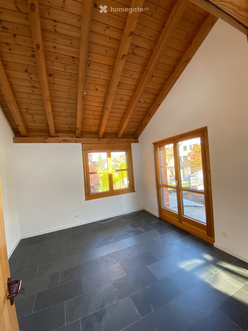 Empty attic room with wooden slatted ceiling, dark grey tiled floor, white walls, and two windows with wooden frames, one of which is a sliding glass door.
