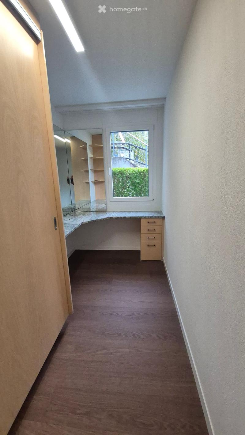 Narrow hallway with carpeted floor, white walls, and a small window showing greenery outside. Features a built-in desk with drawers and a mirror on the left side.