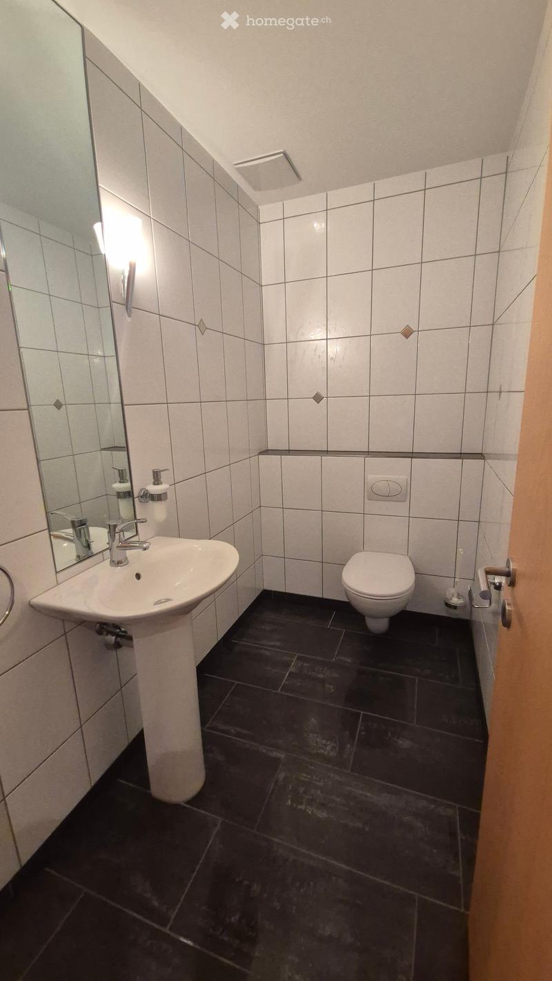 Modern bathroom with white tiled walls, black tiled floor, white sink, chrome faucet, toilet, and mirror. Features a sleek design with a small window near the ceiling.