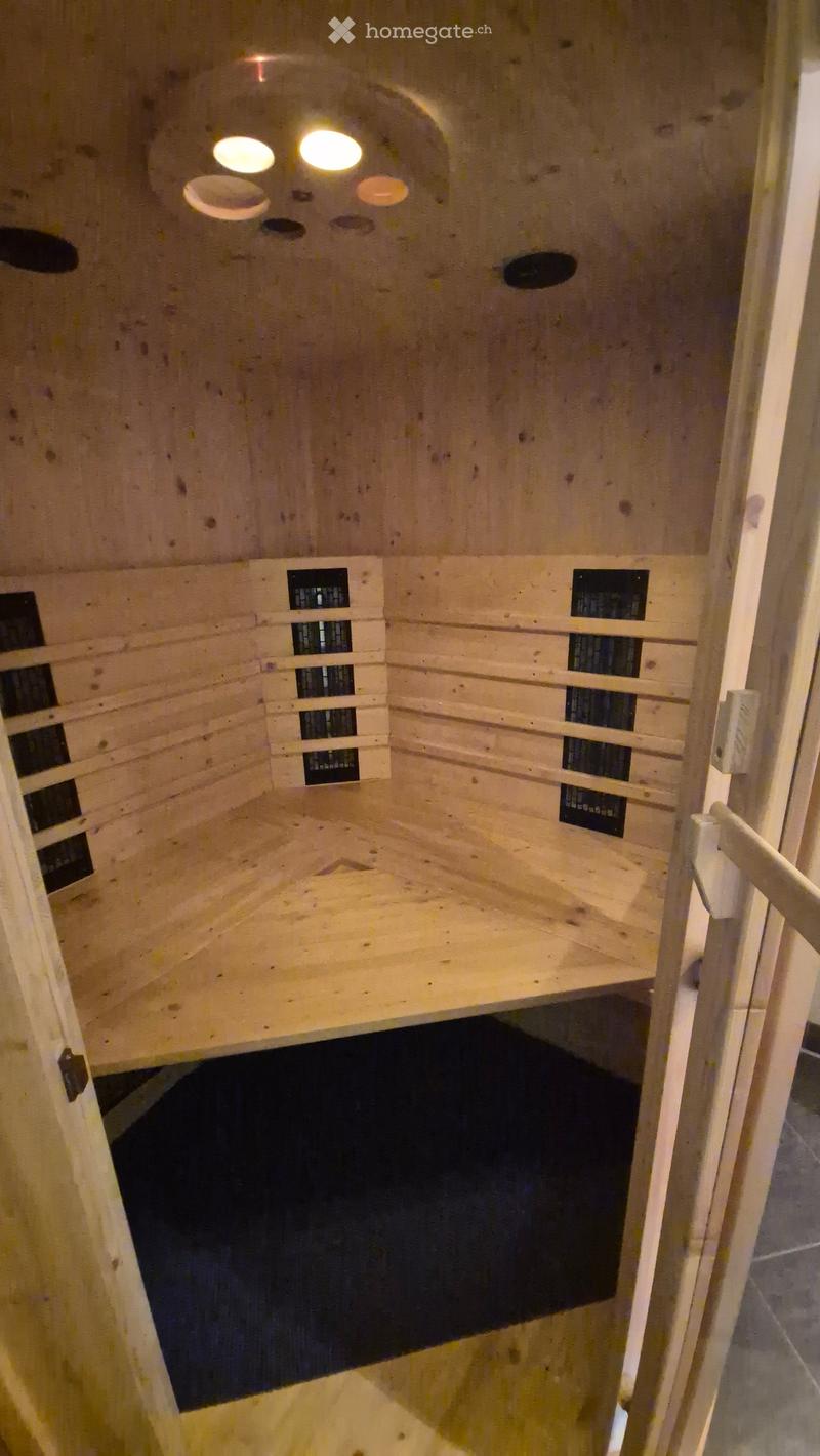 Empty wooden sauna room with built-in benches and a small seating area, featuring a glass door and ceiling lights.