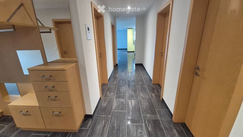 A clean, modern hallway with dark tiled flooring, white walls, and several wooden doors leading to rooms. A small wooden cabinet with drawers is visible on the left side.