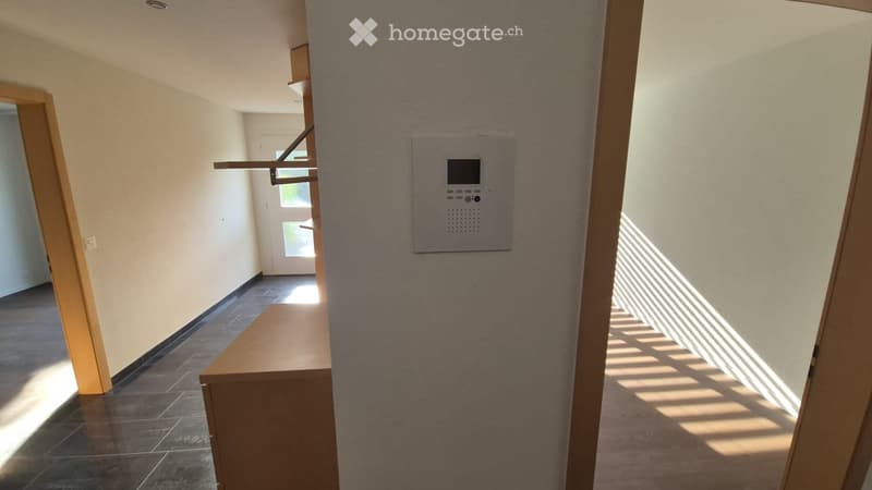 A clean, well-lit hallway with tiled flooring, light-colored walls, and a small wooden bench. A security panel is mounted on the wall, and a door leads to another room.