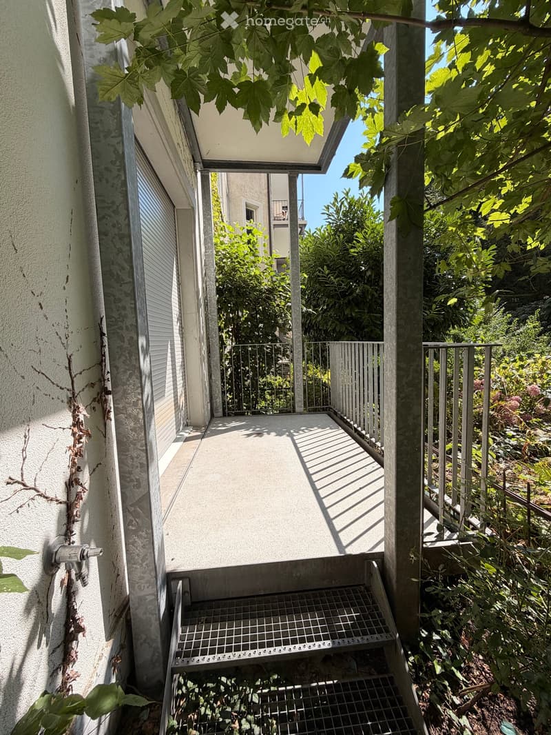 Balcony with metal railing, steps leading down, surrounded by greenery and trees.