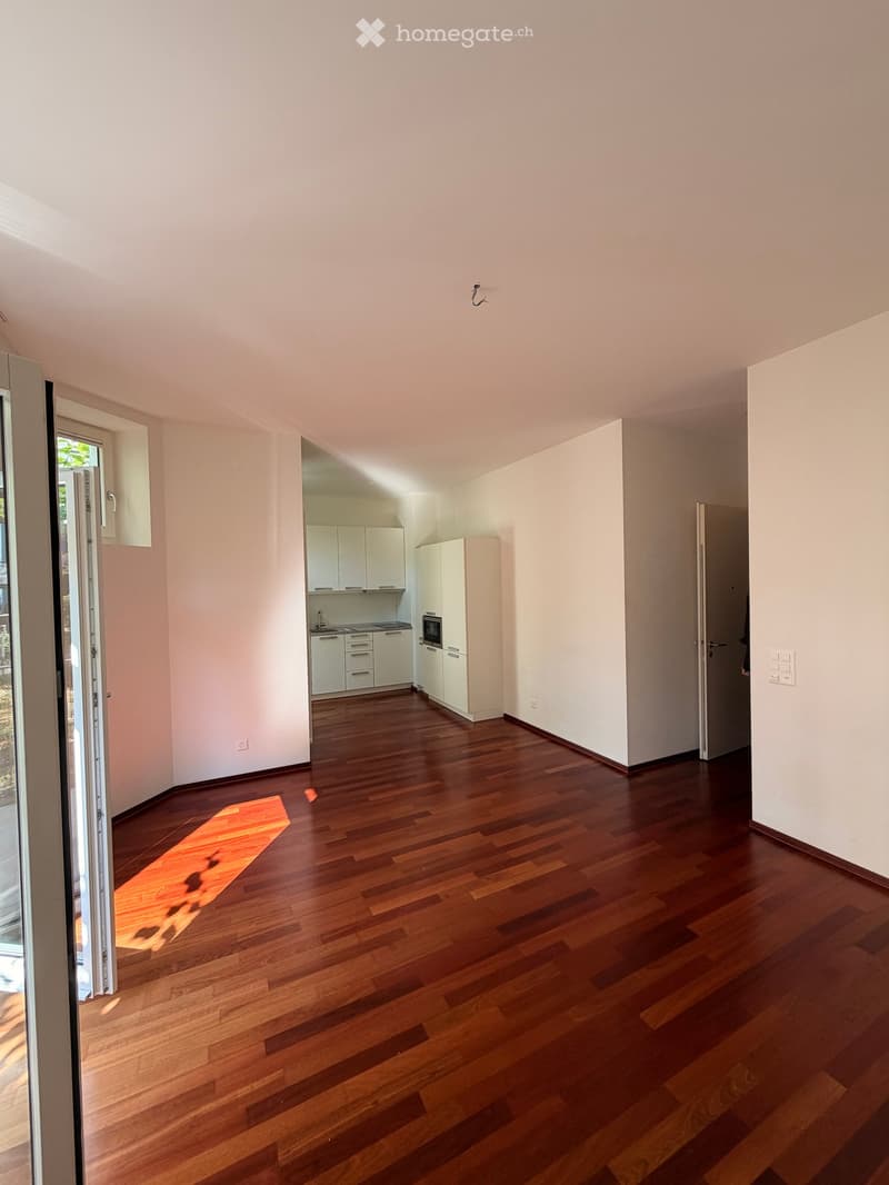 Empty room with wooden flooring, white walls, and a small kitchen area visible in the background.