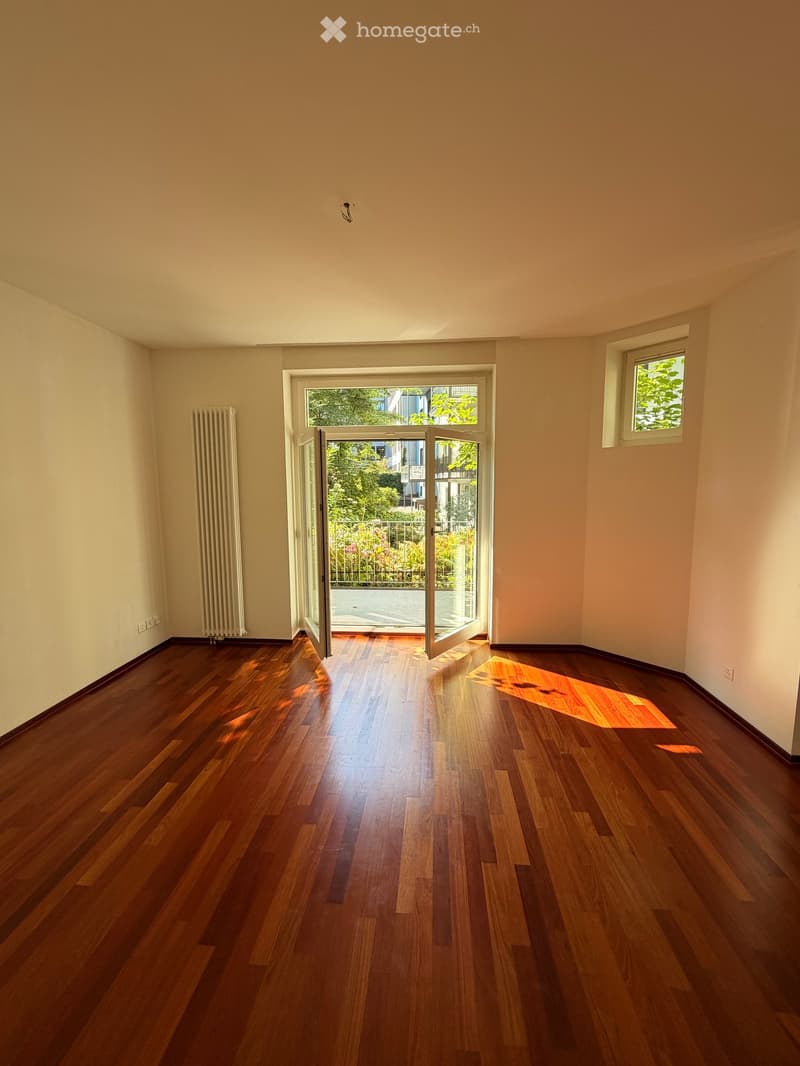 Empty room with wooden flooring, white walls, a radiator, and a large open doorway leading to a garden area.