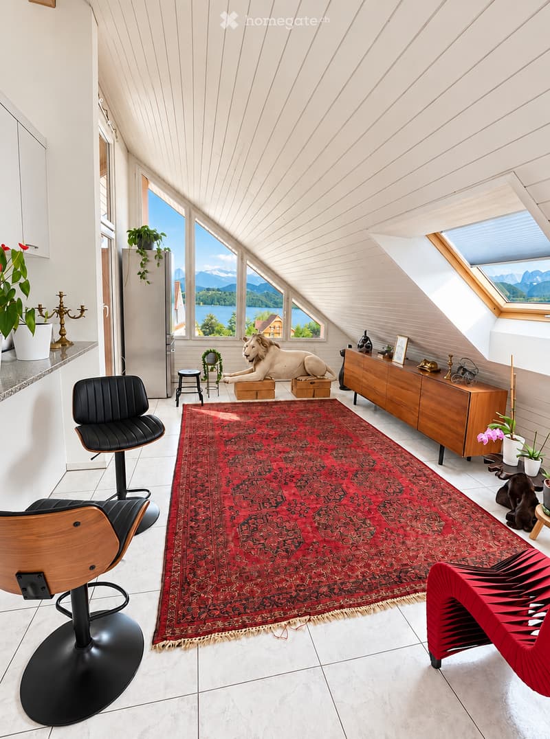 Bright, airy room with slanted wooden ceilings and large windows offering a scenic mountain view. Features include a red patterned rug, a black leather chair, a wooden stool, a wooden cabinet, and decorative items including a lion statue and plants.