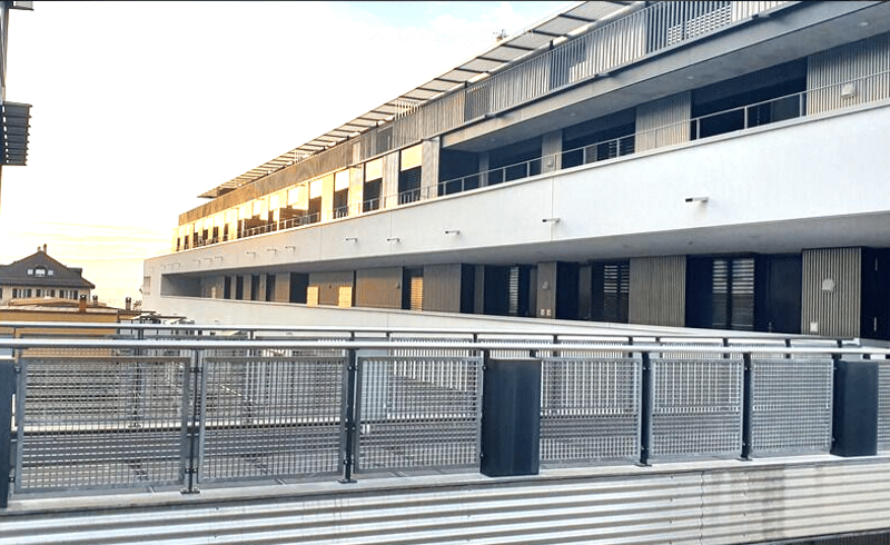 Modern multi-story building with a flat roof and large windows, featuring a metal fence in the foreground.