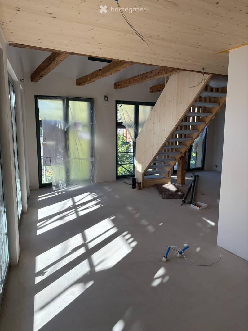 Empty room with exposed wooden beams, a staircase, large windows, and construction materials scattered on the floor.