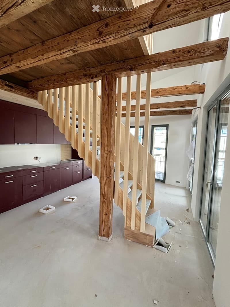 Open-plan space with exposed wooden beams and a wooden staircase under construction. Kitchen area with dark cabinets and countertops is visible on the left. The floor is unfinished concrete, and construction materials are scattered around.