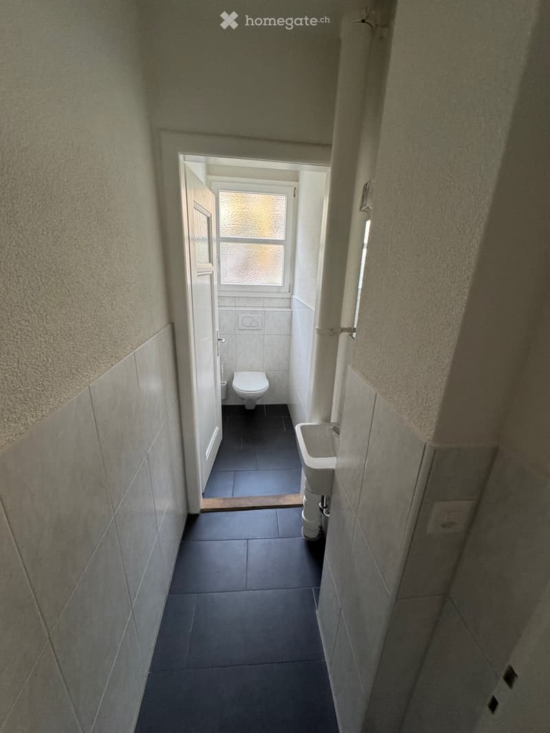 A narrow hallway with tiled walls and floor, leading to a small bathroom with a toilet and sink.