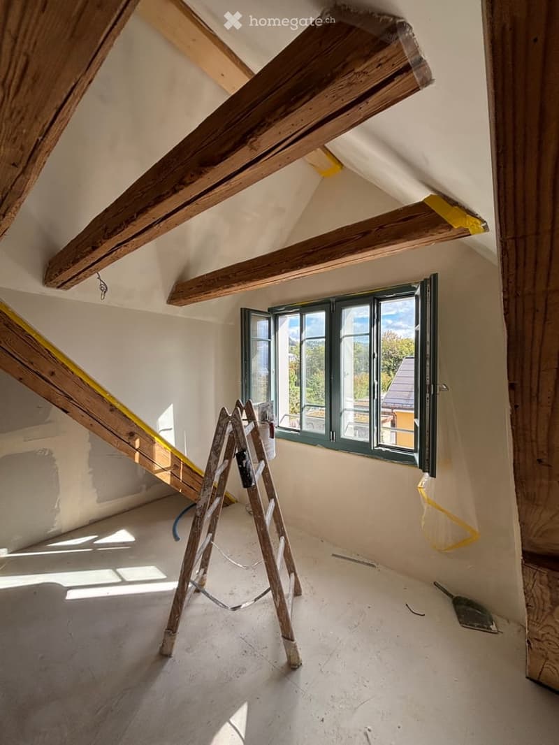 Empty attic space with exposed wooden beams, a window with green frame, a ladder, and construction tools.
