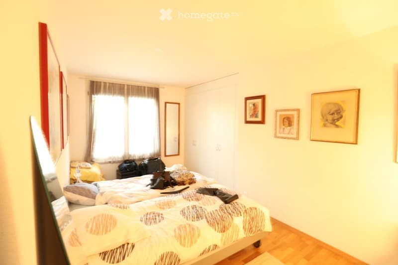 Bright bedroom with a double bed, nightstand, mirror, and several picture frames on the wall. The bed has patterned bedding and is partially made up.
