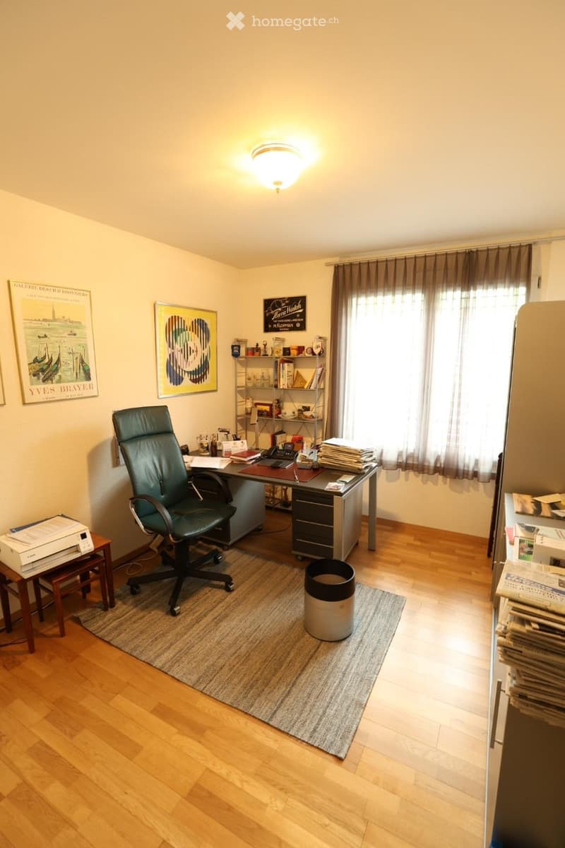 Office space with wooden flooring, a large window with curtains, a desk with a chair, a shelf with books and office supplies, and a small table with a printer.