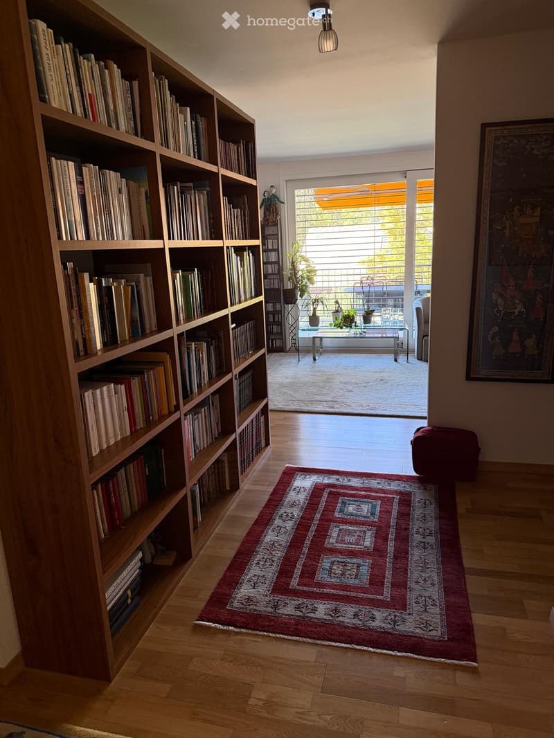 A well-lit hallway with wooden flooring, a large bookshelf filled with books, a patterned red rug, a small table and chairs, and a painting on the wall. The hallway leads to a balcony area with outdoor furniture and plants.