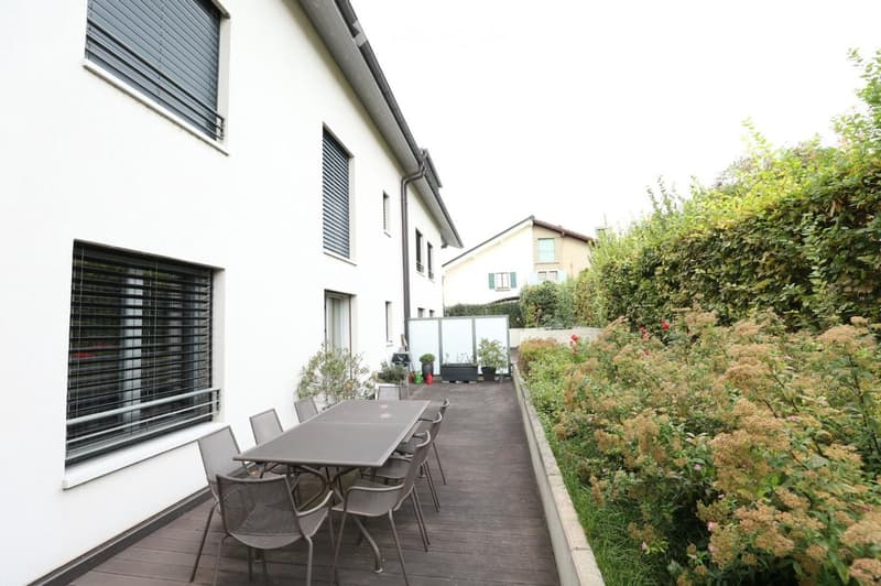 Balcony with wooden decking, grey metal table and chairs, surrounded by greenery and a white building.