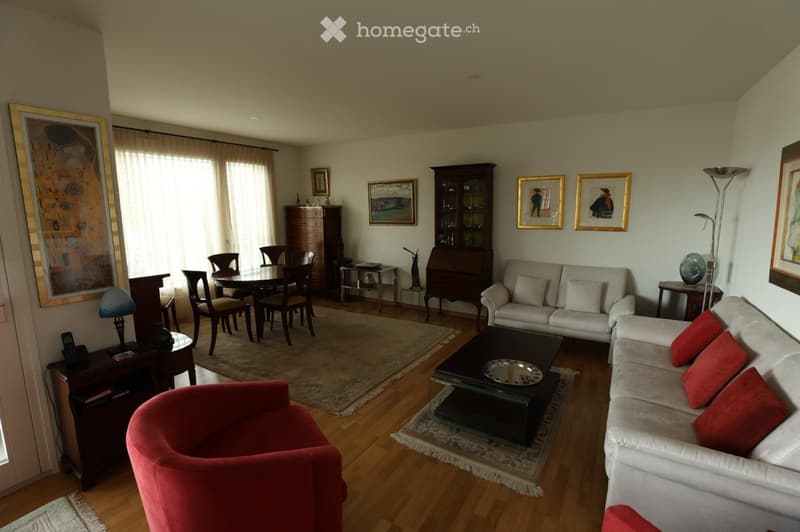 Spacious living room with hardwood floors, large windows, cream-colored sofa with red cushions, coffee table, dining area with table and chairs, side table with lamp, and decorative cabinet.