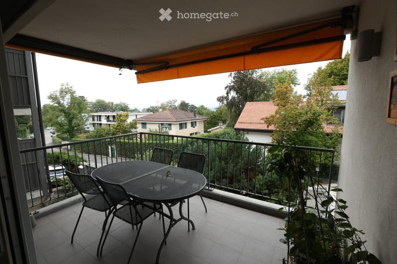 Balcony with a round metal table and four matching chairs, a retractable awning, and a view of a residential area with trees and houses.