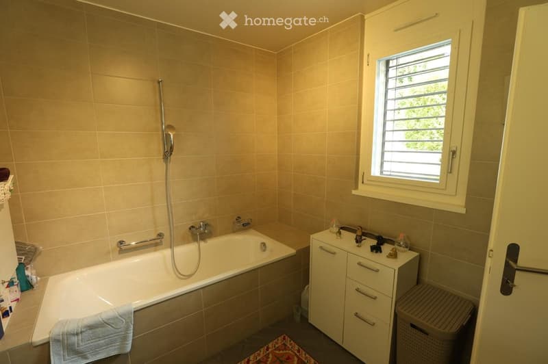 Bathroom with a bathtub, shower hose, faucet, bathroom cabinet, towel, and wicker basket. Features a window with blinds and beige tiled walls.