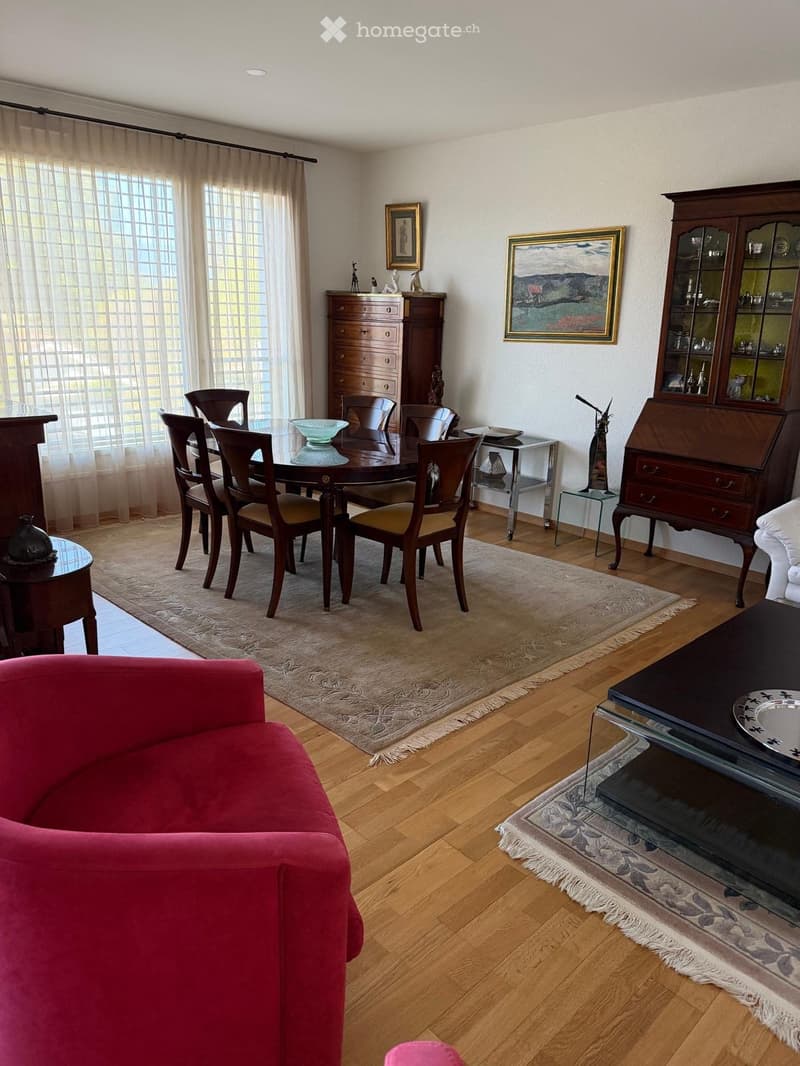 Spacious living and dining area with wooden flooring, large windows with curtains, a red sofa, a dining table with chairs, a coffee table, and decorative cabinets.