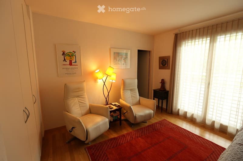 Cozy living room with two white leather armchairs, a small table with a lamp, a red area rug, and large windows with white curtains.
