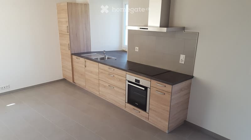 Modern kitchen with light wooden cabinets, black countertop, stainless steel sink, oven, and range hood. Light grey tiled floor.
