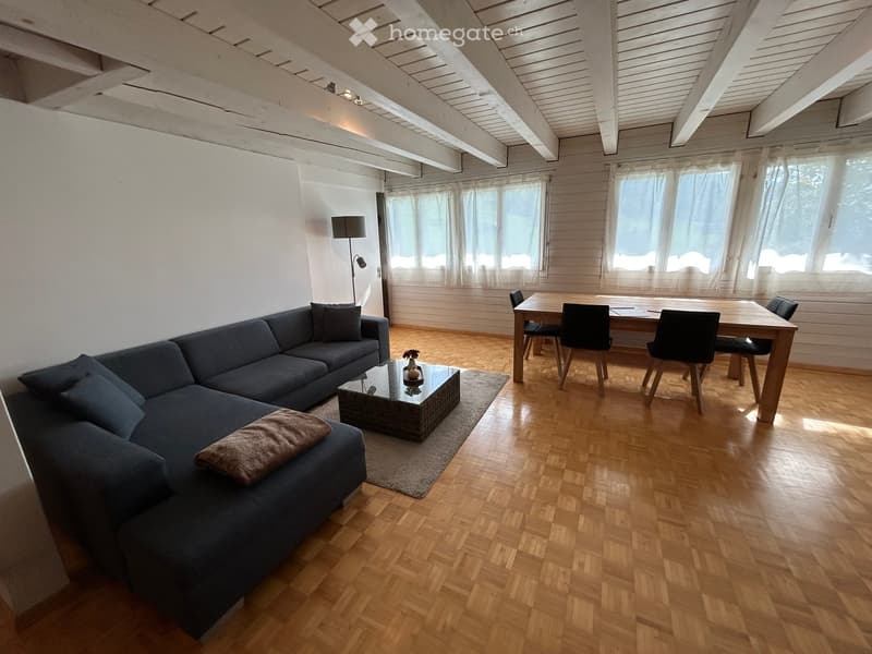 Living room with wooden flooring, exposed wooden ceiling beams, large windows with white curtains, a dark blue sectional sofa, a small coffee table, and a dining area with a wooden table and chairs.