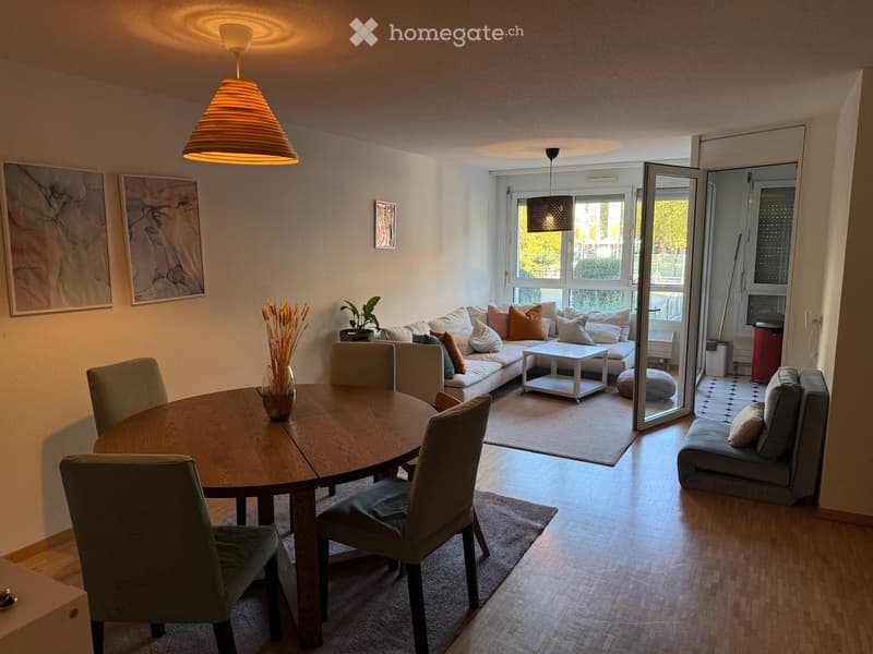 Modern living and dining area with a round wooden dining table, four chairs, a sofa with multiple cushions, a coffee table, a small ottoman, a stool, and a vase with dried flowers on the table. Large windows and a sliding glass door lead to a balcony.