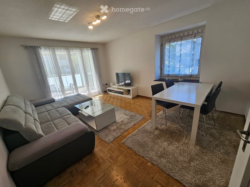 Modern living room with a grey sectional sofa, glass coffee table, white dining table with chairs, and a TV on a white stand. The room features wooden flooring, large windows with curtains, and two area rugs.