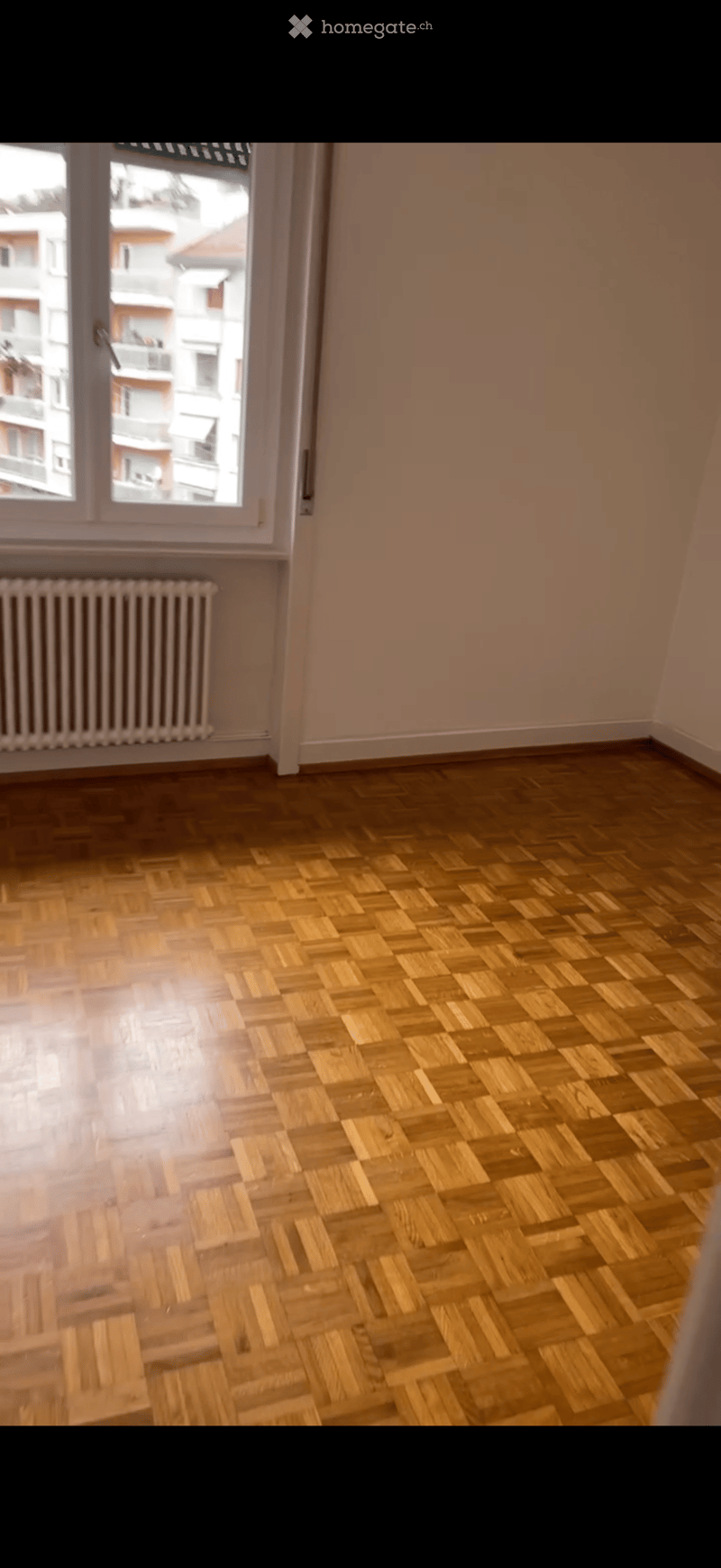 Empty room with wooden herringbone flooring, light-colored walls, and a window with a radiator below it.