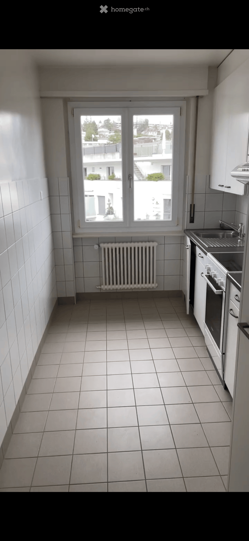 Empty kitchen with tiled floor, white tiled walls, a radiator, a sink, and a window with a view of the building exterior.