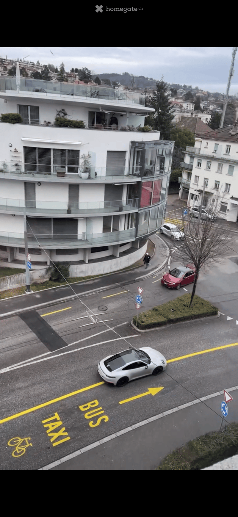 A modern multi-story building with balconies and large windows, surrounded by a street with several parked cars and traffic markings for buses, taxis, and bicycles.