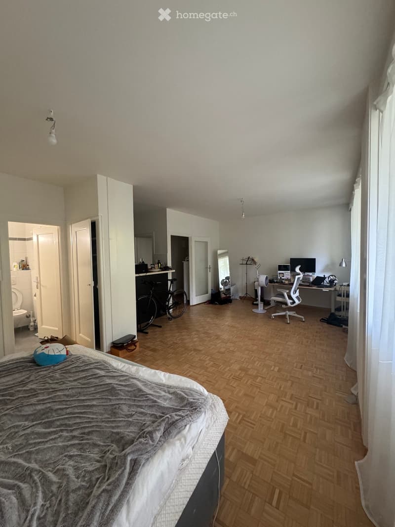 A furnished bedroom with a bed, desk, chair, laptop, mirror, bicycle, and a visible bathroom area with a toilet.
