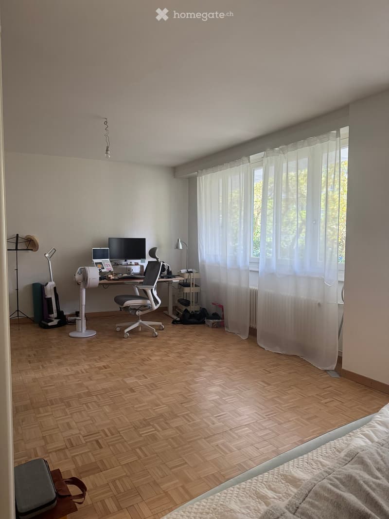 A well-lit room with a wooden herringbone floor, a desk with a computer and chair, a bed with a blanket, and a large window with white curtains.