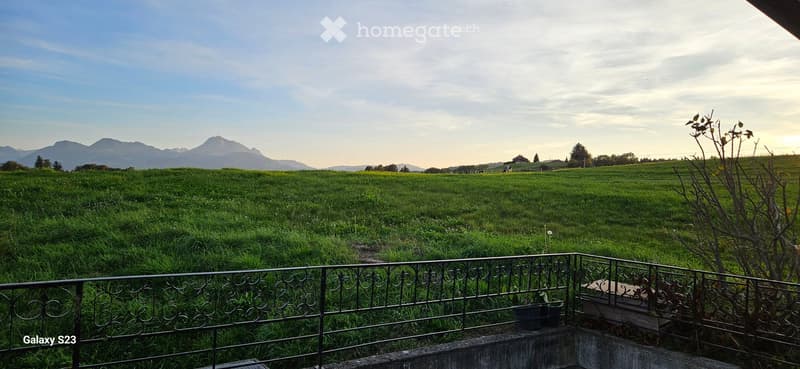 Balcony overlooking a lush green field with distant mountains, featuring a metal railing and a small wooden bench.