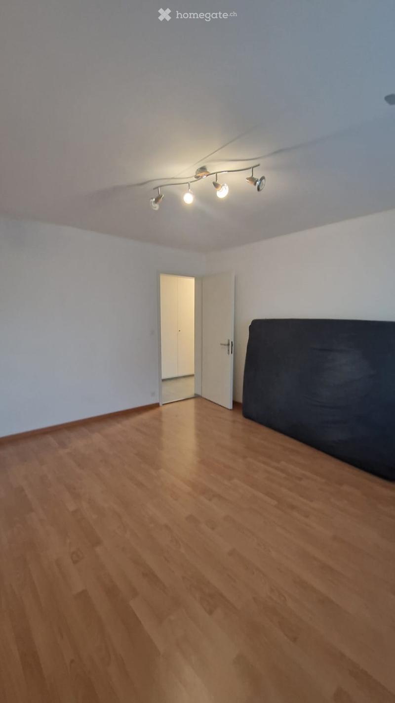 Empty room with wooden flooring, white walls, and a ceiling light fixture. A partially visible mattress is on the right side, and an open door leads to another room.