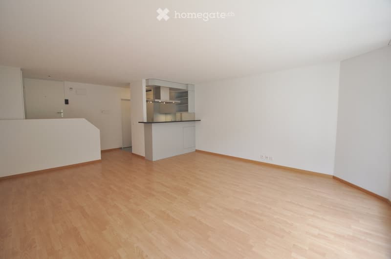 Empty room with light wooden flooring, white walls, and a small kitchen area with cabinets and a countertop.