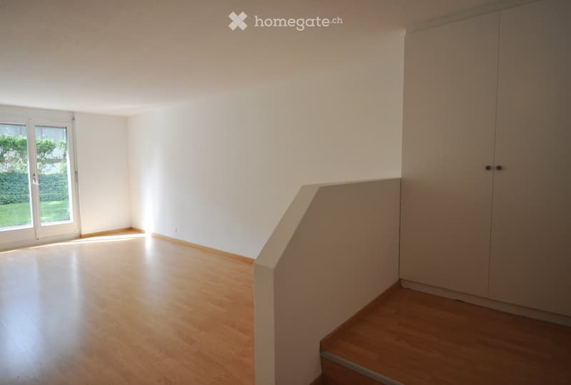 Empty room with wooden flooring, white walls, and a built-in closet. A large window with blinds allows natural light to enter, and a small staircase leads upwards.