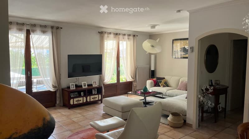 Bright living room with large windows, white sofa, ottoman, coffee table, armchair, TV, and decorative items.