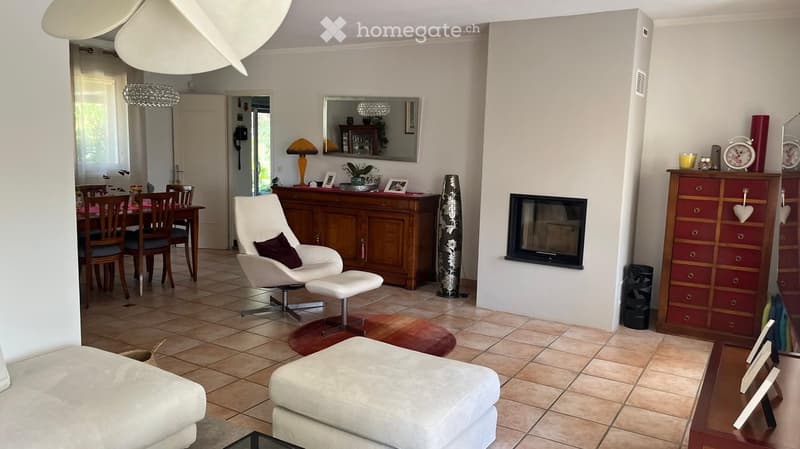 Living room with a white sofa, ottoman, white armchair, and a dining area with a table and chairs. Features a fireplace, a wooden cabinet, and a mirror on the wall.