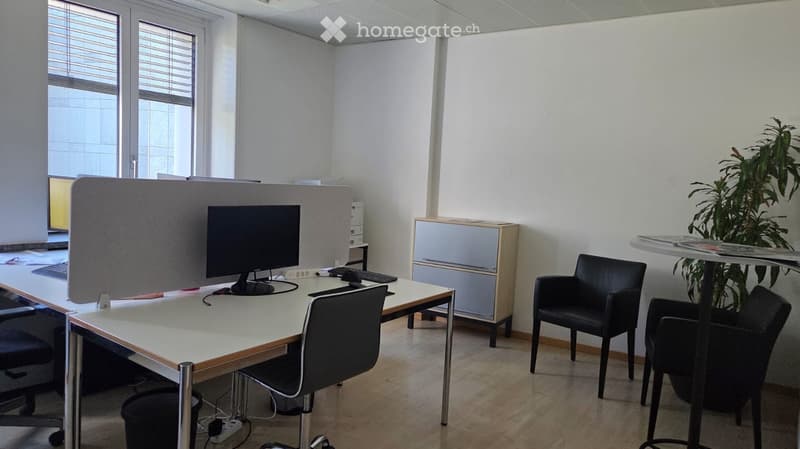 Office space with a desk, computer, chairs, and a cabinet. Large windows with blinds provide natural light.