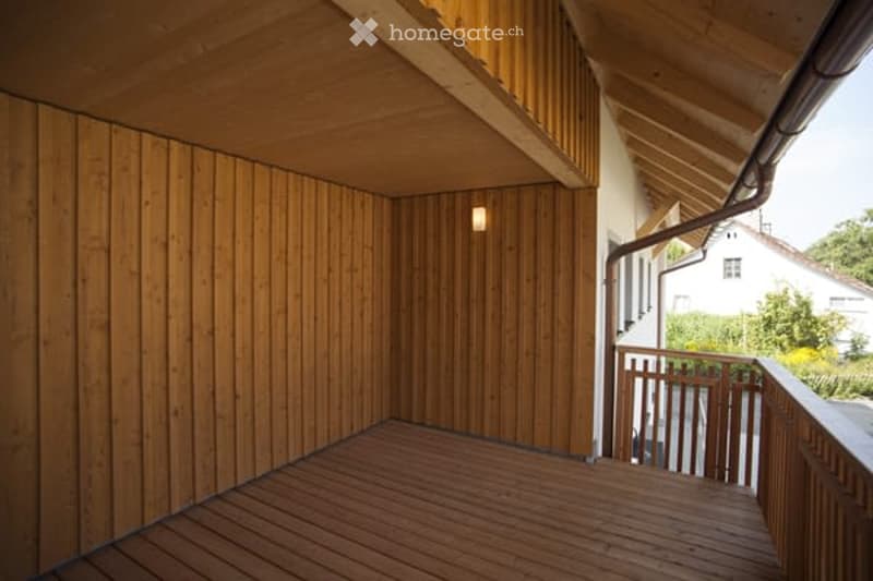 Wooden balcony with slatted wooden walls and ceiling, small light fixture, open railing, and view of greenery and neighboring house.