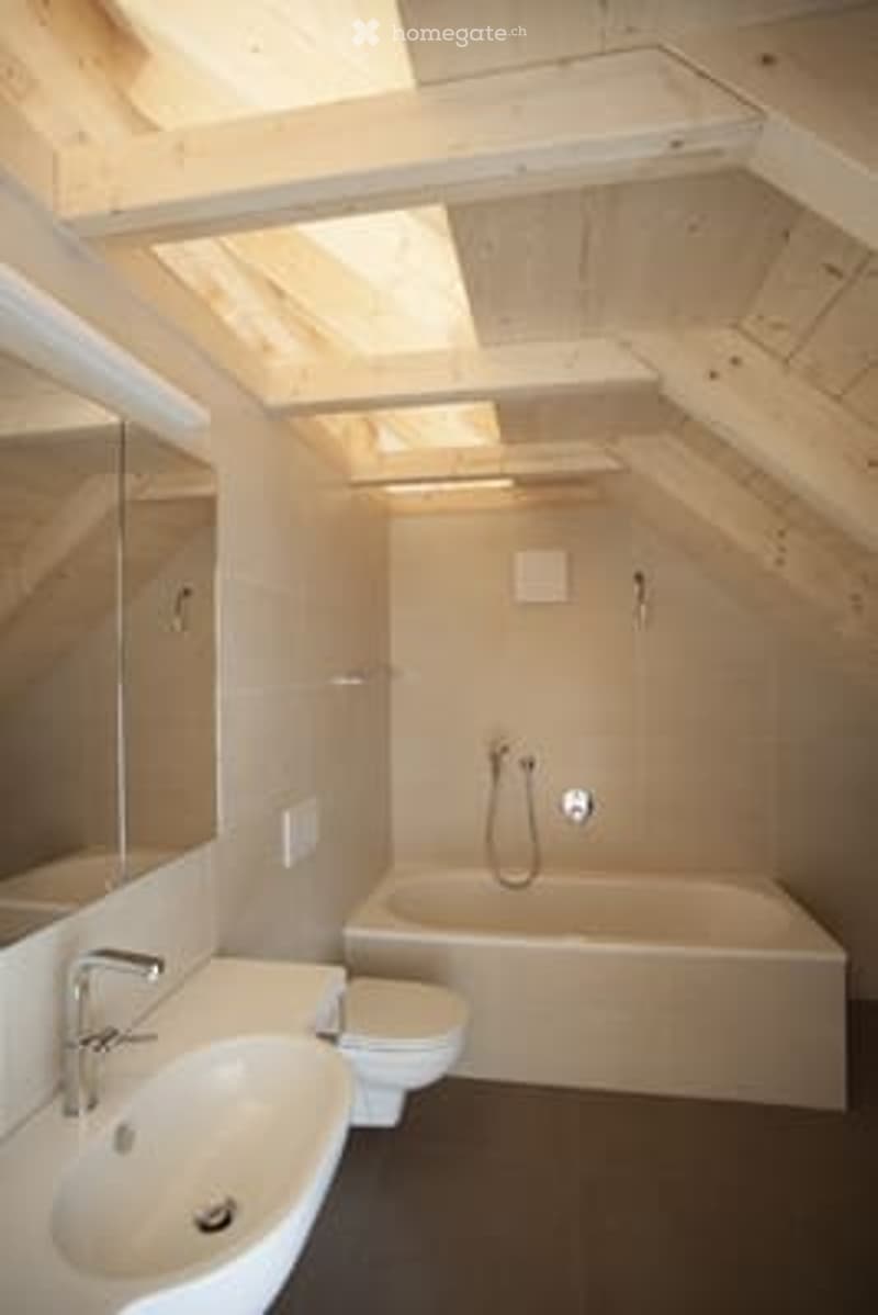 Modern bathroom with wooden slanted ceiling, large mirror, sink, toilet, bathtub, and showerhead.
