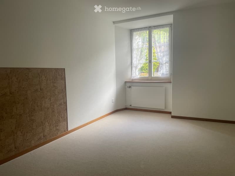 Empty room with light-colored walls, carpeted floor, and a single window with white curtains. A radiator is mounted below the window.